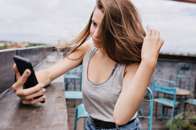 Hermosa modelo femenina en ropa casual tomando una foto de sí misma en la cafetería de la azotea. Foto al aire libre de una impresionante dama jugando con su cabello mientras hace selfie en un día frío de verano.