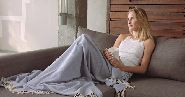 Hermosa modelo femenina en un sofá con una taza de café bajo una manta azul mirando a la cámara y luego volviendo a sus pensamientos