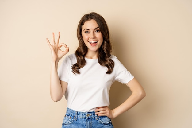 Hermosa modelo femenina sonriendo complacida, mostrando bien, signo cero, halagando, alabándote, de pie sobre un fondo beige.