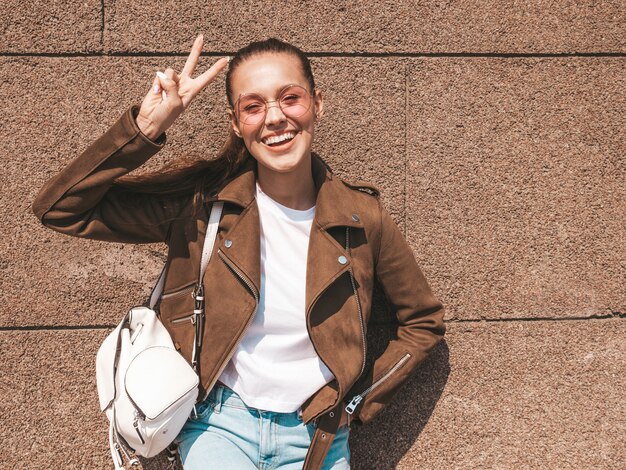 Hermosa modelo morena sonriente vestida con chaqueta hipster de verano y ropa de jeans mostrando el signo de la paz