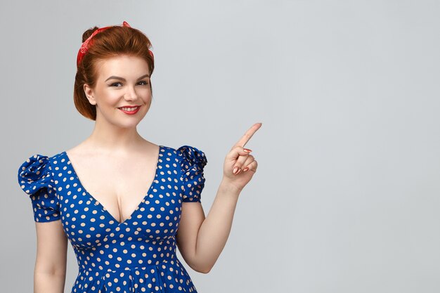 Hermosa modelo de mujer joven positiva con peinado elegante y maquillaje glamoroso mirando a la cámara con una encantadora sonrisa feliz, apuntando con el dedo índice a la pared vacía con espacio de copia para su anuncio