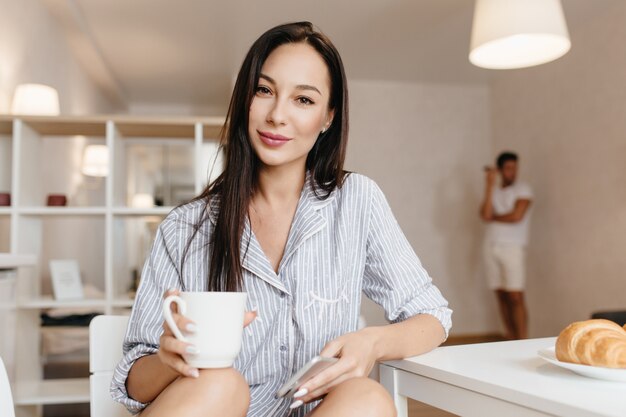 Hermosa modelo de mujer morena en camisa azul posando en la cocina con una taza de café