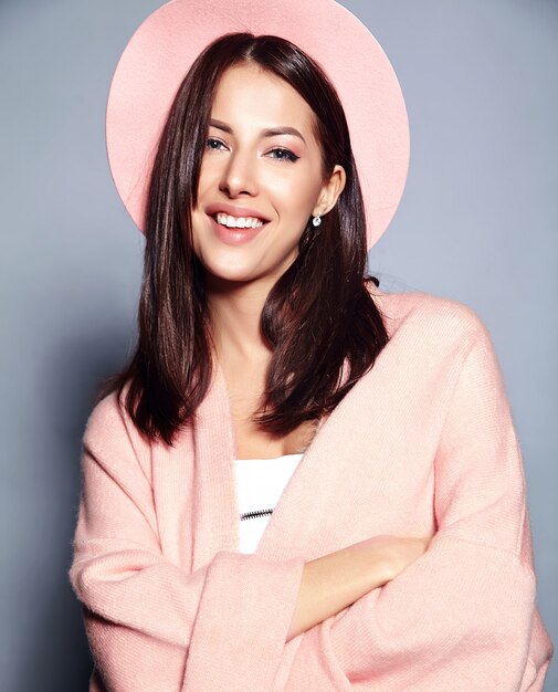 Hermosa modelo de mujer morena hipster sonriente en elegante abrigo rosa y colorido sombrero posando en gris