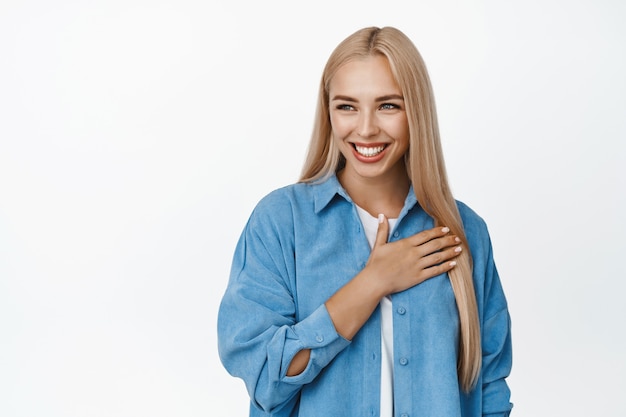 Hermosa modelo de mujer sosteniendo la mano en el corazón y sonriendo, mirando a la izquierda en el espacio de la copia, gesto de agradecimiento, de pie en blanco