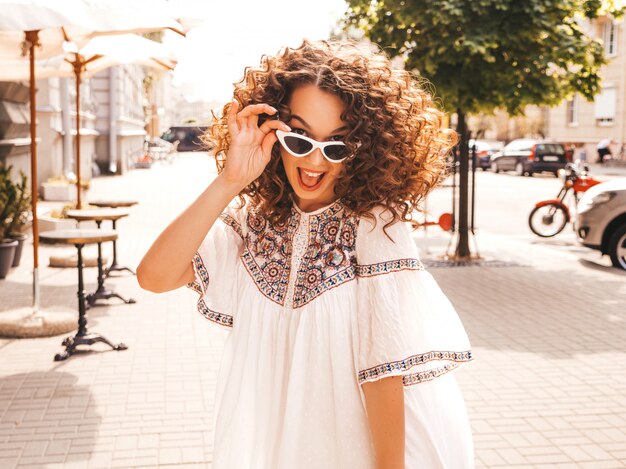 Hermosa modelo sonriente con peinado afro rizos vestido con vestido blanco hipster de verano.