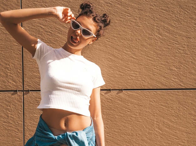 Hermosa modelo sonriente con peinado de cuernos vestido con ropa de camiseta blanca hipster de verano. Chica despreocupada sexy posando en la calle cerca de la pared. Moda mujer divertida y positiva divirtiéndose en gafas de sol