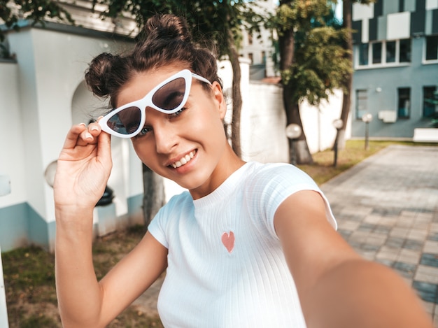 Hermosa modelo sonriente con peinado de cuernos vestido con ropa casual de verano. Chica despreocupada sexy posando en la calle con gafas de sol. Tomando fotos de autorretrato selfie en teléfono inteligente