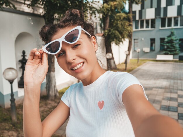 Hermosa modelo sonriente con peinado de cuernos vestido con ropa casual de verano. Chica despreocupada sexy posando en la calle con gafas de sol. Tomando fotos de autorretrato selfie en teléfono inteligente