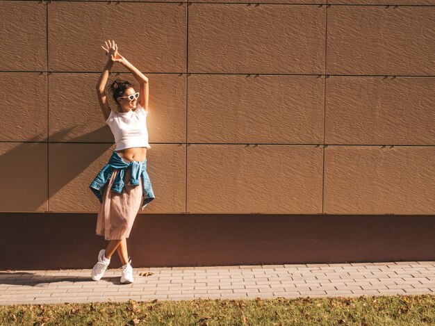 Hermosa modelo sonriente con peinado de cuernos vestido con ropa de jeans de chaqueta hipster de verano. Chica despreocupada sexy posando en la calle cerca de la pared. Moda mujer divertida y positiva divirtiéndose en gafas de sol