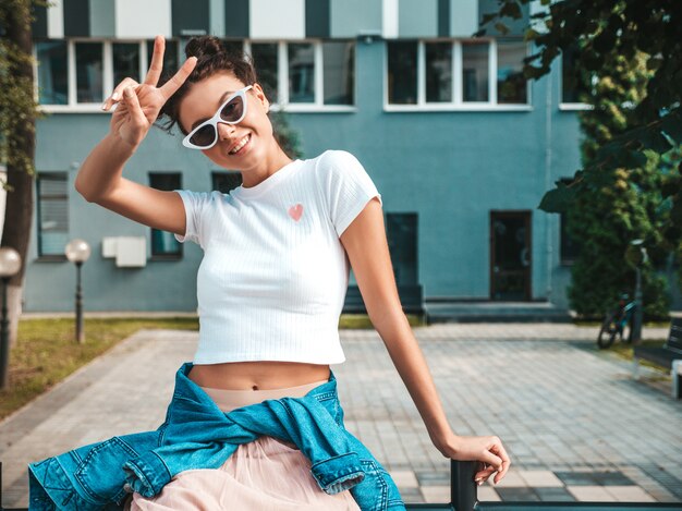 Hermosa modelo sonriente con peinado de cuernos vestido con ropa de jeans de chaqueta hipster de verano. Chica despreocupada sexy posando en la calle. Mujer divertida y positiva de moda divirtiéndose en gafas de sol