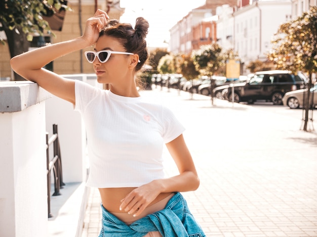 Hermosa modelo sonriente con peinado de cuernos vestido con ropa de jeans de chaqueta hipster de verano. Chica despreocupada sexy posando en la calle. Mujer divertida y positiva de moda divirtiéndose en gafas de sol