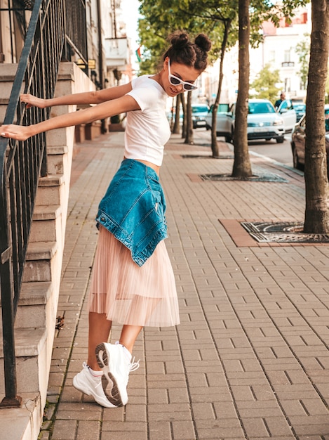 Hermosa modelo sonriente con peinado de cuernos vestido con ropa de jeans de chaqueta hipster de verano. Chica despreocupada sexy posando en la calle. Mujer divertida y positiva de moda divirtiéndose en gafas de sol