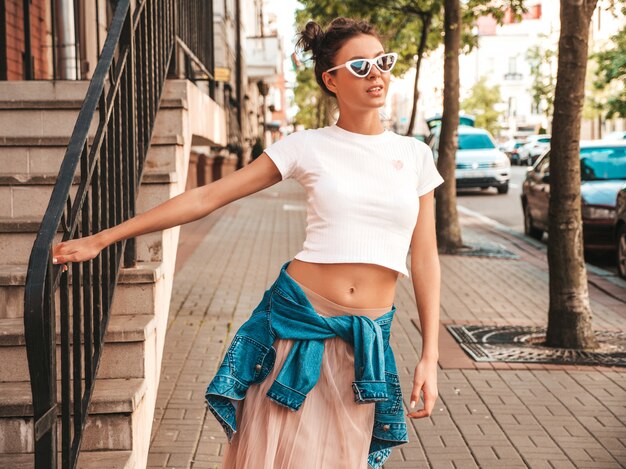 Hermosa modelo sonriente con peinado de cuernos vestido con ropa de jeans de chaqueta hipster de verano. Chica despreocupada sexy posando en la calle. Mujer divertida y positiva de moda divirtiéndose en gafas de sol