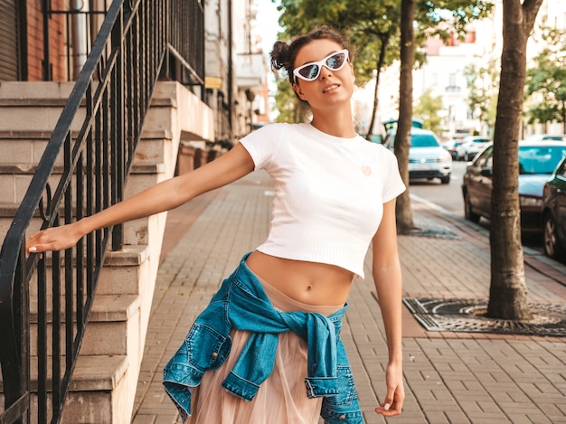 Hermosa modelo sonriente con peinado de cuernos vestido con ropa de jeans de chaqueta hipster de verano. Chica despreocupada sexy posando en la calle. Mujer divertida y positiva de moda divirtiéndose en gafas de sol