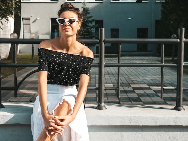 Hermosa modelo sonriente vestida con ropa elegante de verano. Chica despreocupada sexy sentada en la calle. Empresaria moderna moderna en gafas de sol divirtiéndose