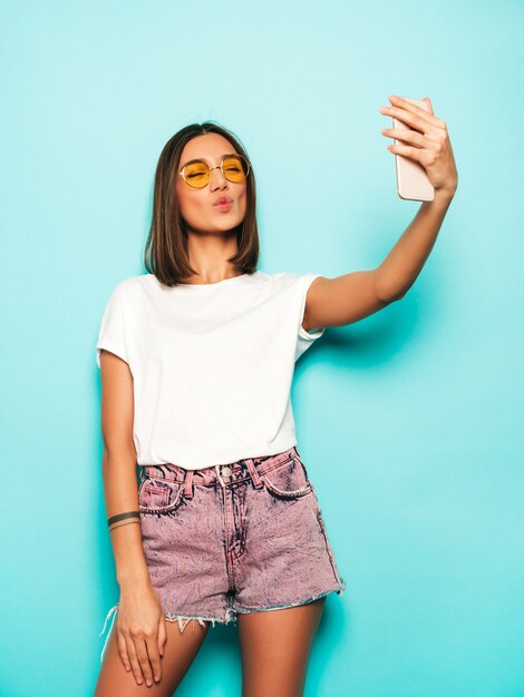 Hermosa modelo sonriente vestida con ropa hipster de verano. Chica despreocupada sexy posando en estudio junto a la pared azul en shorts de jeans. Mujer de moda y divertida tomando fotos de autorretrato selfie en teléfono inteligente