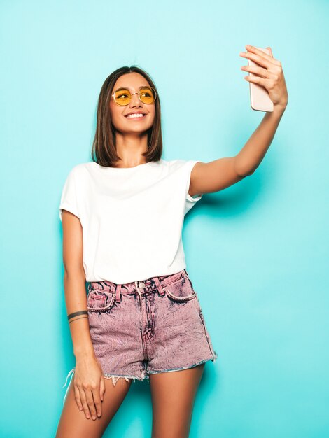 Hermosa modelo sonriente vestida con ropa hipster de verano. Chica despreocupada sexy posando en estudio junto a la pared azul en shorts de jeans. Mujer de moda y divertida tomando fotos de autorretrato selfie en teléfono inteligente