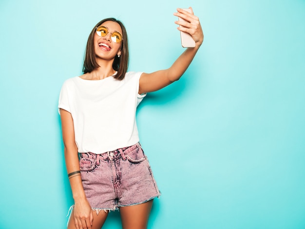 Hermosa modelo sonriente vestida con ropa hipster de verano. chica despreocupada sexy posando en estudio junto a la pared azul en shorts de jeans. mujer de moda y divertida tomando fotos de autorretrato selfie en teléfono inteligente