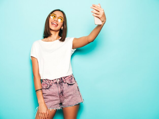 Hermosa modelo sonriente vestida con ropa hipster de verano. Chica despreocupada sexy posando en estudio junto a la pared azul en shorts de jeans. Mujer de moda y divertida tomando fotos de autorretrato selfie en teléfono inteligente