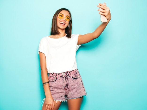 Hermosa modelo sonriente vestida con ropa hipster de verano. Chica despreocupada sexy posando en estudio junto a la pared azul en shorts de jeans. Mujer de moda y divertida tomando fotos de autorretrato selfie en teléfono inteligente