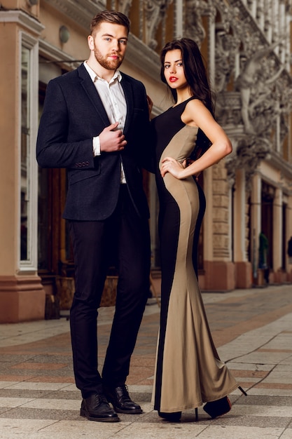 Hermosa morena elegante con su marido caminando por las calles de la ciudad de ale. Disfrutando de su tiempo, vistiendo traje clásico negro y vestido de cóctel largo.