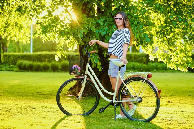 Hermosa morena se encuentra en el césped verde con bicicleta en el parque