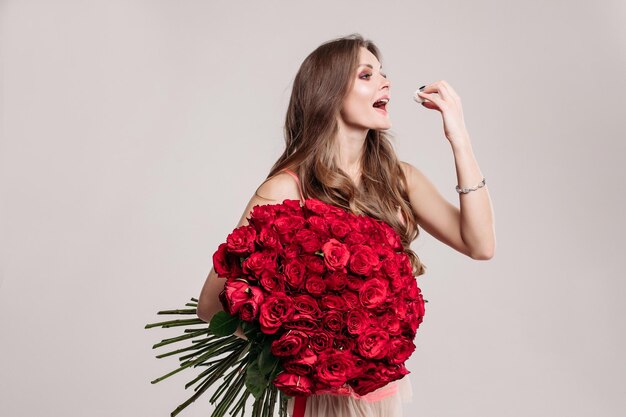 Hermosa morena feliz con el pelo largo y ondulado sosteniendo un hermoso ramo de rosas rojas y comiendo un delicioso postre con la mano Aislada en el fondo blanco del estudio