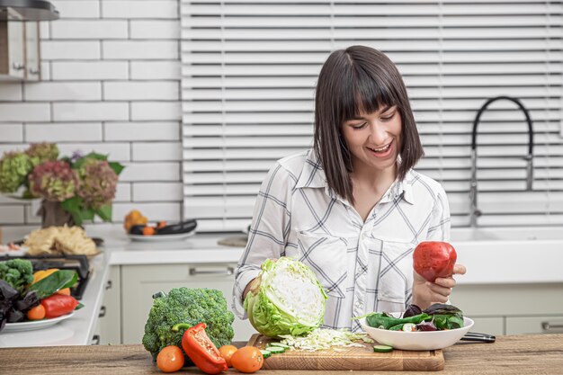 Hermosa morena sonríe y corta verduras en una ensalada en el fondo de un interior de cocina moderna.