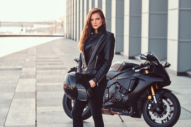 Una hermosa motociclista con chaqueta de cuero negro sosteniendo casco al lado de una superbike fuera de un edificio.