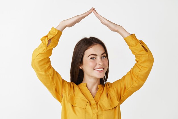 Hermosa mujer adulta haciendo un techo de la casa con las manos por encima de la cabeza, sonriendo y mirando feliz, de pie en la pared blanca