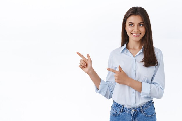 Hermosa mujer adulta joven en blusa azul presenta la pancarta de su compañía, sonriendo amable y feliz, apuntando hacia la izquierda en el espacio en blanco para su anuncio, pared de estudio de pie