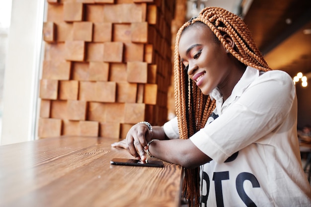 Hermosa mujer africana con elegante camisa casual y rastas posando en el café cerca del alféizar de la ventana y tocando su teléfono celular