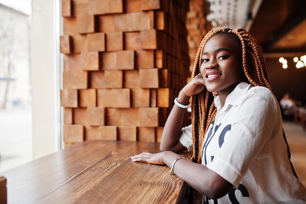 Hermosa mujer africana con elegante camisa casual y rastas posando en el café cerca del alféizar de la ventana