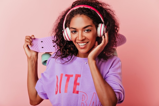 Hermosa mujer africana con patineta sonriendo. Retrato interior de una romántica chica rizada en auriculares aislados en rosa.