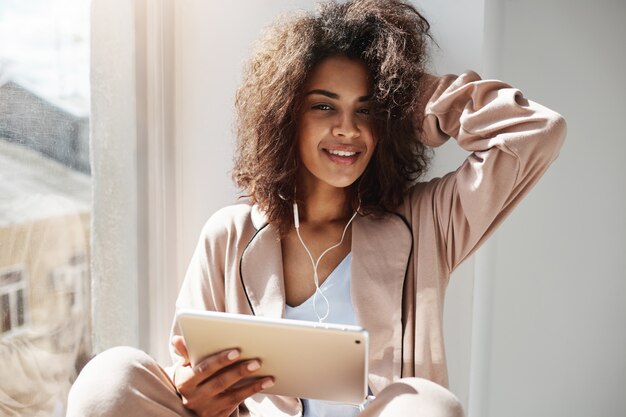 Hermosa mujer africana en ropa de dormir y auriculares sonriendo sosteniendo tableta sentado en el alféizar de la ventana escuchando música en streaming por la mañana.