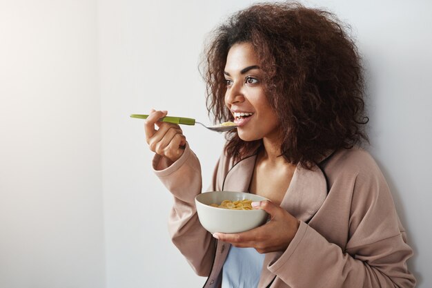 Hermosa mujer africana en ropa de dormir sonriendo comiendo copos con leche en casa.