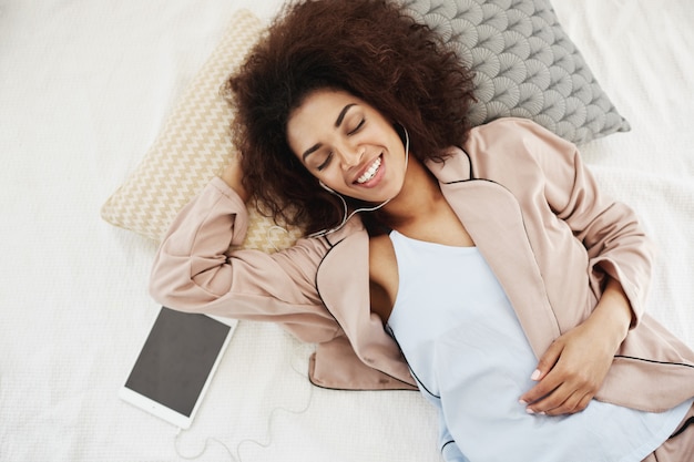 Hermosa mujer africana en ropa de dormir sonriendo escuchando música en los auriculares acostado en la cama.