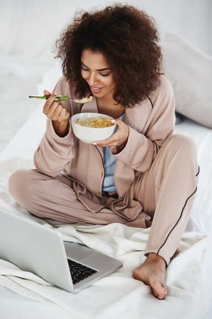 Hermosa mujer africana en ropa de dormir sonriendo mirando portátil comiendo copos con leche sentado en la cama.