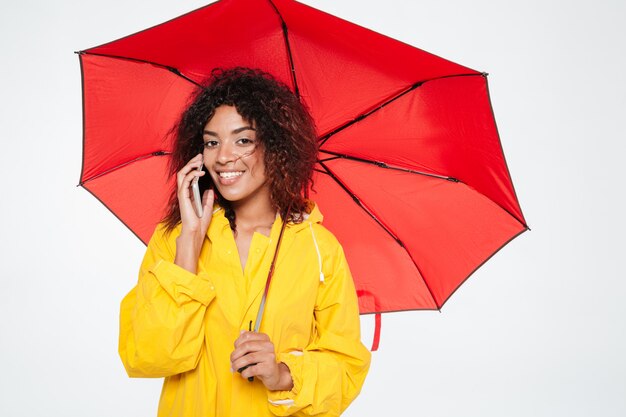 Hermosa mujer africana sonriente en gabardina escondiéndose bajo el paraguas mientras habla por teléfono inteligente y mirando a la cámara sobre fondo blanco.