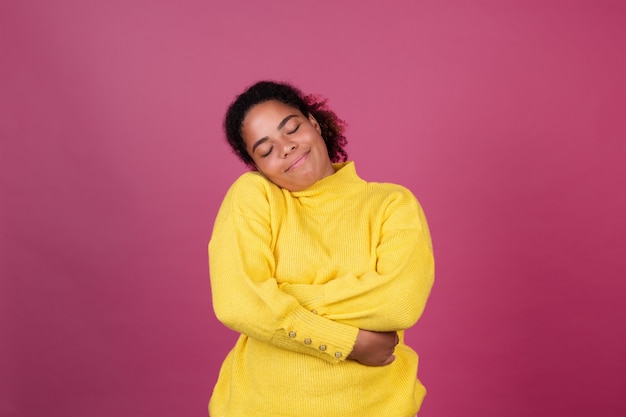 Hermosa mujer afroamericana en pared rosa feliz sonriendo abrazándose a sí misma, ámate a ti mismo concepto, cuidado personal