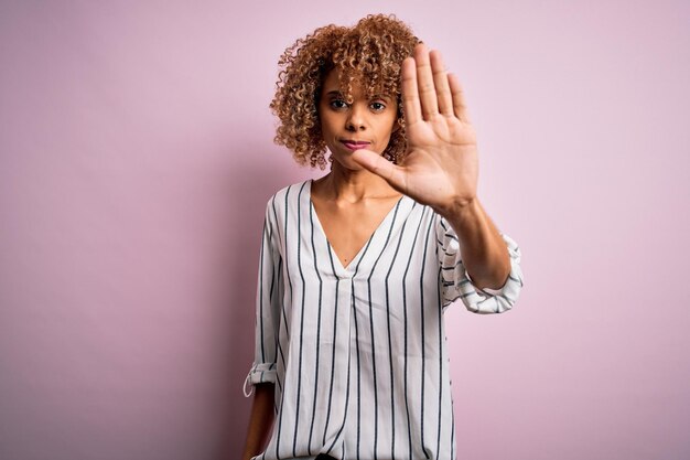 Hermosa mujer afroamericana con el pelo rizado y camiseta a rayas sobre fondo rosa dejando de cantar con la palma de la mano Expresión de advertencia con gesto negativo y serio en la cara