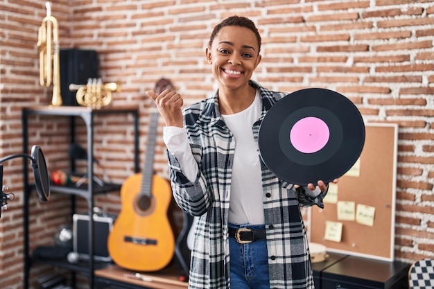 Foto gratuita hermosa mujer afroamericana sosteniendo un disco de vinilo en el estudio de música apuntando con el pulgar hacia el lado sonriendo feliz con la boca abierta