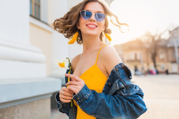 Hermosa mujer agitando el pelo sonriendo, ropa elegante, con chaqueta vaquera y top amarillo, tendencia de moda, estilo de verano, estado de ánimo positivo feliz, día soleado, amanecer, moda callejera, gafas de sol azules