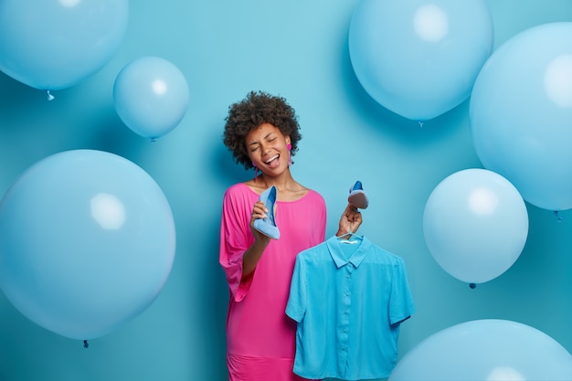 Hermosa mujer alegre con cabello afro elige atuendo para una cita romántica, se jacta de ropa nueva y zapatos comprados a la venta en una tienda de ropa, canta sin preocupaciones, sostiene la camisa en perchas, aislado en azul