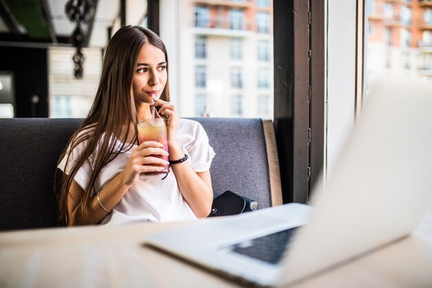 Hermosa mujer alegre freelancer de buen humor, usando una computadora portátil, sentado en cafés, bebiendo Mojitos.
