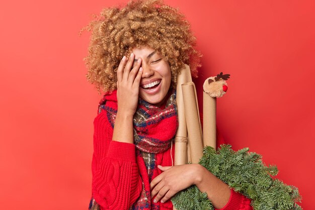 Hermosa mujer alegre hace que la cara de la palma se ría felizmente mantiene los ojos cerrados usa suéter y bufanda alrededor del cuello lleva papel enrollado de corona de abeto verde aislado sobre fondo rojo. Decoraciones