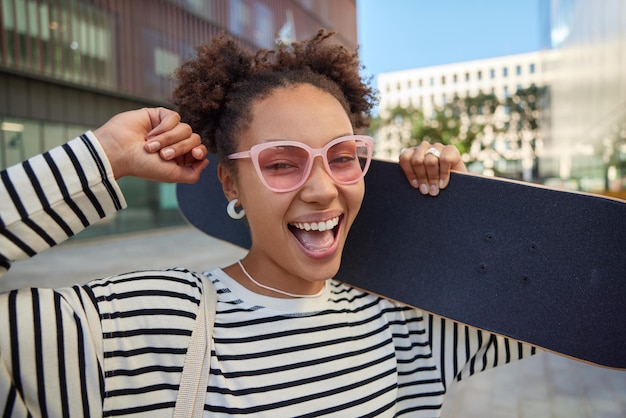 Foto gratuita hermosa mujer alegre con el pelo rizado lleva el monopatín en el hombro se ríe felizmente pasea durante el día soleado en la ciudad