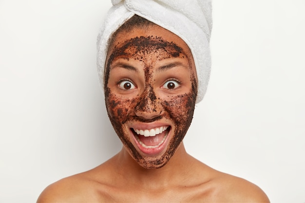 Hermosa mujer alegre con una piel fresca y saludable, sonríe ampliamente, se ve con una reacción feliz de sorpresa, se aplica una mascarilla facial exfoliante de café para reducir los puntos oscuros en la piel, tiene terapia de spa después de la ducha