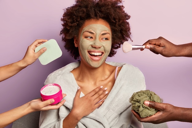 Hermosa mujer alegre renovada con mascarilla de arcilla nutritiva mira felizmente a un lado, siendo tratada con crema, esponjas y pincel de maquillaje