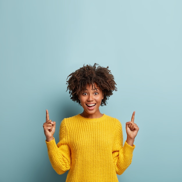 Hermosa mujer alegre y sonriente con peinado afro, apunta hacia arriba, muestra una promoción genial u oferta increíble, vestida con un suéter amarillo, da consejos, posa sobre fondo azul